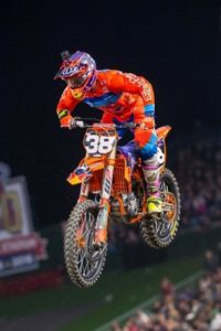 Ein Fahrer in orangefarbener KTM-Motorradbekleidung springt während eines Motocross-Events durch die Luft.