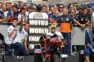Teamfeier des Moto3-Weltmeisters 2022 mit jubelnden Teammitgliedern und Motorrädern auf dem Rennplatz.