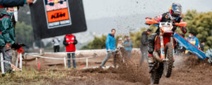 Ein KTM Motorradfahrer springt auf einer Motocross-Strecke und wirbelt dabei Staub auf, während Zuschauer im Hintergrund beobachten.