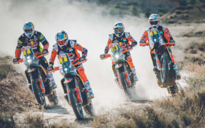 Vier KTM-Motorräder fahren in einer staubigen Landschaft, während die Fahrer in orangefarbener Rennbekleidung mit Helmen und Schutzkleidung die Strecke meistern.