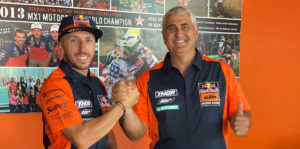Zwei Männer in KTM-Kleidung lächeln und schütteln sich die Hände vor einer Wand mit Motorsportmotiven.